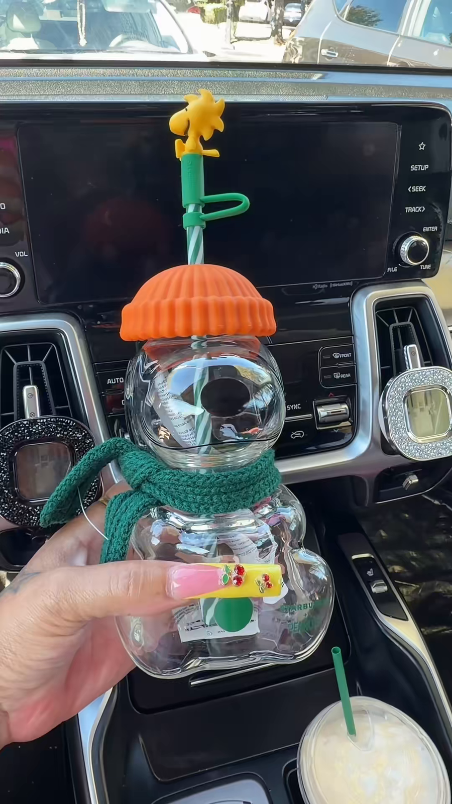 Snoopy glass cup with an orange lid and green scarf held in front of a car dashboard.