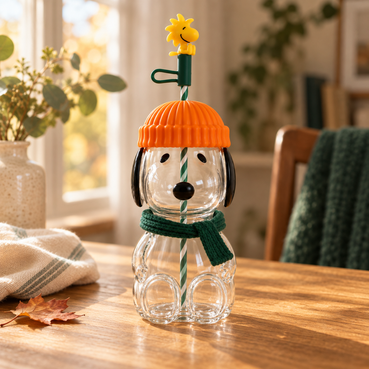 Transparent Pup-kin glass cup with an orange lid and green scarf on a sunlit wooden table.